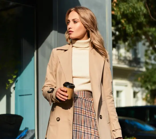 Elegancka kobieta w beżowym płaszczu, kremowym golfie i spódnicy w kratę, z kubkiem kawy w dłoni. Inspiracja na stylizacje do beżowego płaszcza.